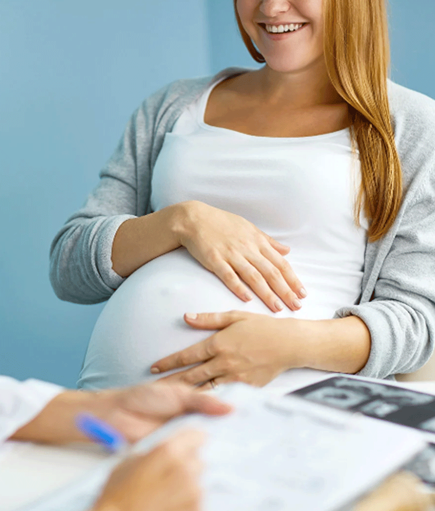 A imagem captura um momento de cuidado pré-natal, mostrando uma mulher grávida sorridente e tocando sua barriga, sugerindo que ela está conversando ou tendo uma consulta com um profissional de saúde, cujo foco está em um documento ou formulário, provavelmente relacionado à sua gravidez. Embora a imagem não mostre explicitamente um cenário odontológico, a cena pode representar uma consulta de pré-natal odontológico, onde o bem-estar oral da futura mãe é discutido e planejado, uma vez que a saúde bucal durante a gravidez é essencial tanto para a mãe quanto para a saúde do bebê. A alegria e expectativa da mãe são evidentes, refletindo a importância de cuidados abrangentes durante este período especial.