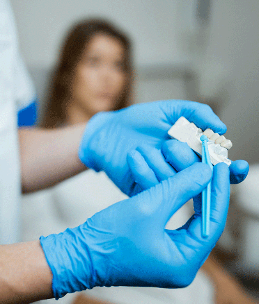 A imagem mostra um profissional de saúde, possivelmente um dentista ou técnico de prótese dentária, usando luvas azuis e segurando um modelo de gesso de dentes humanos. O foco da fotografia está nas mãos do profissional e no modelo dental, que é frequentemente usado para planejamento de tratamento ou criação de próteses dentárias como coroas ou pontes. Ao fundo, desfocado, há uma paciente olhando em direção ao profissional, sugerindo que ela pode estar aguardando um procedimento ou consulta odontológica. A imagem tem uma atmosfera clínica e pode ser usada em materiais educativos sobre procedimentos dentários ou em promoção de serviços odontológicos.